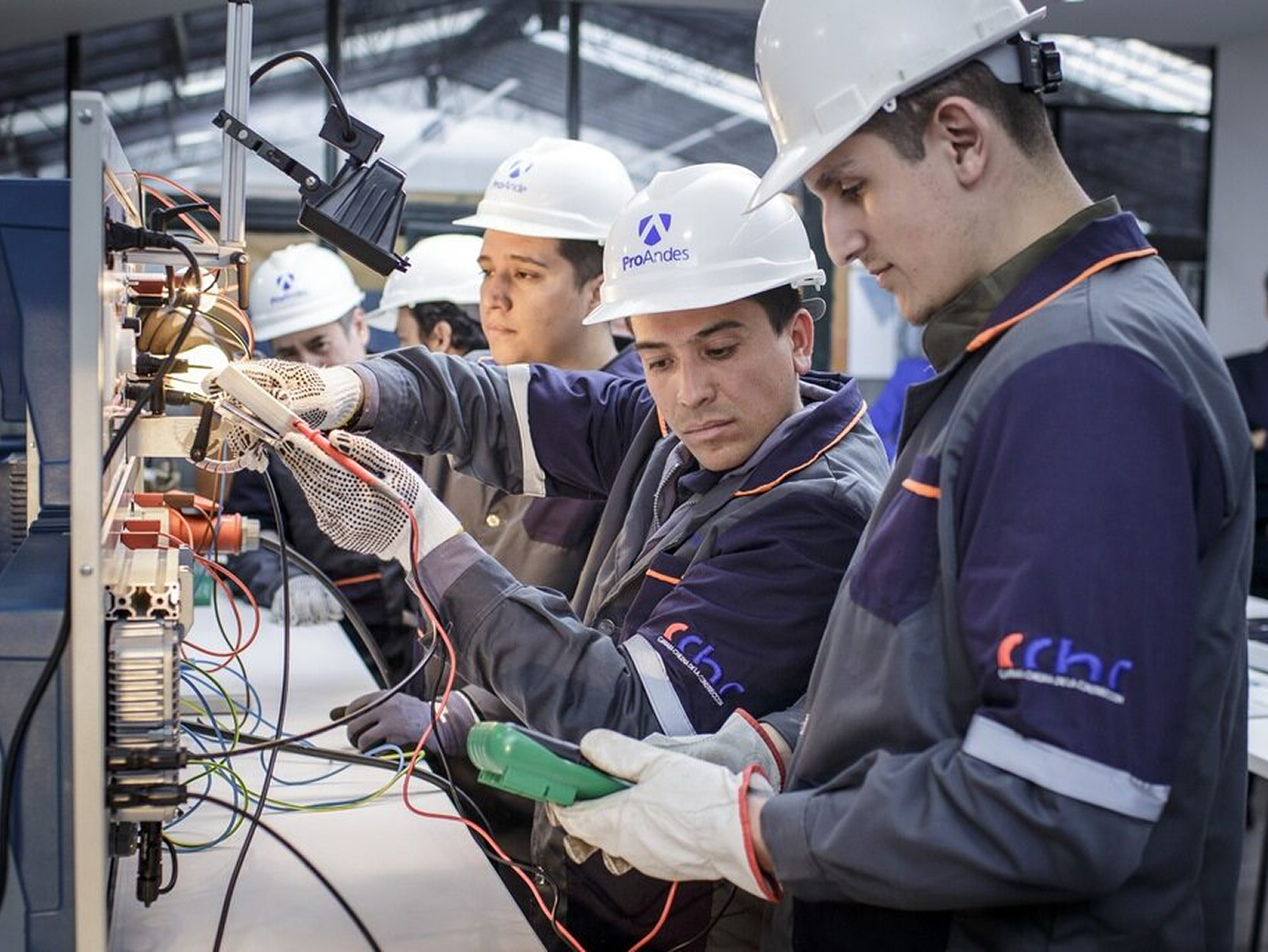 Mehrere Männer in Schutzkleidung und Helmen arbeiten konzentriert an einer elektrischen Schaltung in einer Werkstatt.